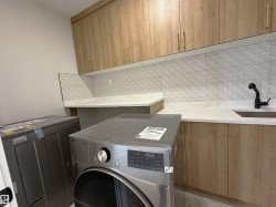 Laundry area featuring wood-finish cabinetry, white countertops, textured backsplash tiling, and a built-in sink with a matte black faucet - 