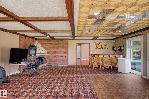 Spacious recreation room featuring a brick fireplace with a metal hood, exposed wood ceiling beams, a built-in bar, and direct access to the outdoors via a glass paned door - 250 197 Avenue, Edmonton, AB - Indoor