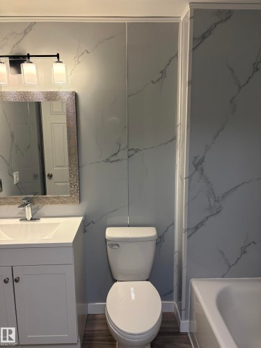 Bathroom featuring marble-look wall panels, a white vanity with a rectangular sink, wood-finish flooring, a contemporary three-light fixture, and a framed mirror - 14075 30 Street E, Edmonton, AB - Indoor Photo Showing Bathroom
