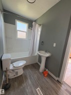 Bathroom featuring a white pedestal sink, white toilet, white bathtub with a shower kit, and wood-finish flooring - 