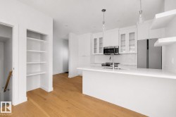 Modern kitchen featuring a white island with an integrated sink, stainless steel appliances, white cabinetry with some glass-front doors, and white floating shelves - 