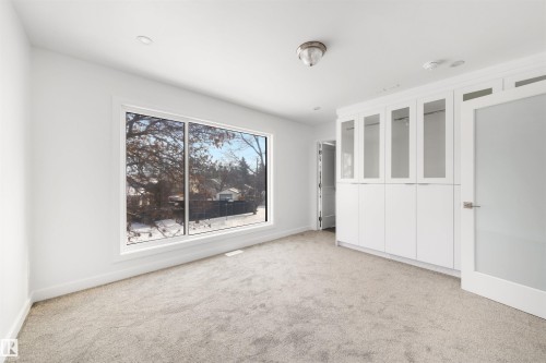 This room features a large window, light-colored carpeting, and a built-in white storage unit with glass-fronted upper cabinets - 7927 112S Avenue, Edmonton, AB - Indoor Photo Showing Other Room