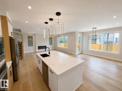 Open-concept living area featuring a large kitchen island with a white countertop and undermount sink, pendant lighting, wood-finish flooring, a linear electric fireplace, and expansive windows - 