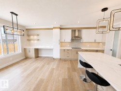 Open-concept kitchen and dining area featuring light wood-finish flooring, a kitchen island with a waterfall countertop, white and wood-tone cabinetry, a stainless steel range hood, and integrated desk space with floating shelving - 