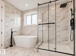 Spacious bathroom featuring a freestanding soaking tub with a matte black floor-mounted faucet, a walk-in shower with a glass enclosure and matte black rain showerhead, and white marble-look tile walls and flooring - 