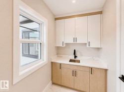 Modern utility space featuring a single-hung window, light wood-finish lower cabinetry, white upper cabinetry, a stainless steel sink, and a black gooseneck faucet - 