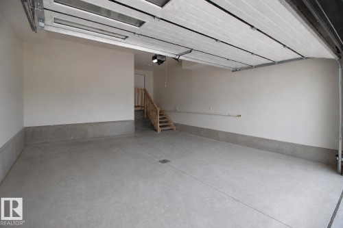Spacious garage featuring a concrete floor, light-toned walls, a partially open sectional garage door with window panels, a built-in wooden staircase, and an overhead garage door opener - 16 Middleton Close, Leduc, AB 