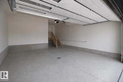 Spacious garage featuring a concrete floor, light-toned walls, a partially open sectional garage door with window panels, a built-in wooden staircase, and an overhead garage door opener - 