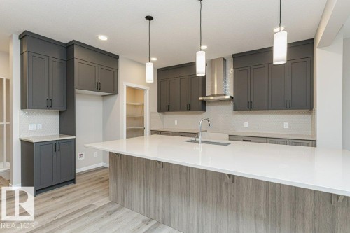 Contemporary kitchen featuring a large central island with a white countertop and wood-finish paneling, dark gray shaker-style cabinetry, a hexagon tile backsplash, and wood-finish flooring - 2616 158 Street, Edmonton, AB - Indoor Photo Showing Kitchen With Upgraded Kitchen