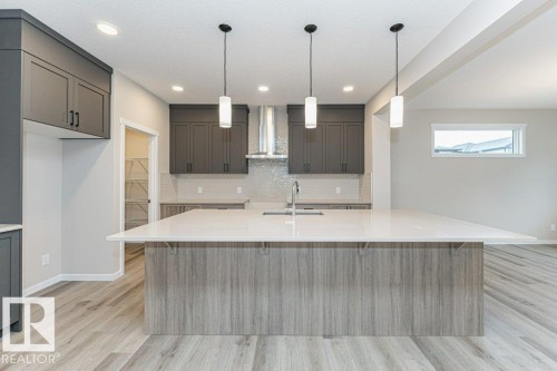 Kitchen featuring a large central island with a light-toned countertop, grey cabinetry, stainless steel range hood, mosaic tile backsplash, and wood-finish flooring - 2616 158 Street, Edmonton, AB - Indoor Photo Showing Kitchen With Upgraded Kitchen