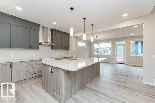 Open-concept kitchen featuring an oversized island with countertop seating, stainless steel range hood, grey upper cabinetry, wood-finish lower cabinetry, and light wood-finish flooring - 2616 158 Street, Edmonton, AB - Indoor Photo Showing Kitchen With Upgraded Kitchen
