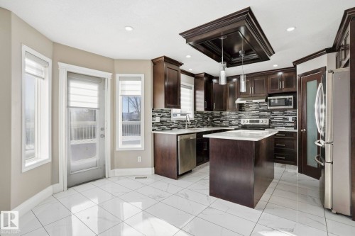 The kitchen features dark wood cabinetry, stainless steel appliances, an island with a light-colored countertop, and a tiled backsplash - 4831 154 Avenue, Edmonton, AB - Indoor Photo Showing Kitchen With Upgraded Kitchen