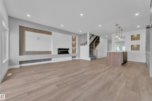 Open-concept living space featuring wood-finish flooring, a contemporary fireplace with a decorative slat wall, a kitchen island with a white countertop and brass pendant lighting, and a staircase with dark carpeting and wood railing - 3116 8 Avenue, Edmonton, AB - Indoor Photo Showing Other Room