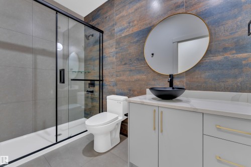 Bathroom featuring a walk-in shower with black-framed glass enclosure, a white toilet, a light gray vanity with a vessel sink, a gold-framed circular mirror, and textured wall tiling - 3116 8 Avenue, Edmonton, AB - Indoor Photo Showing Bathroom