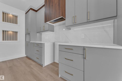 Contemporary kitchen featuring light gray cabinetry with gold-finish hardware, a wood-finish range hood, and white countertops - 3116 8 Avenue, Edmonton, AB - Indoor