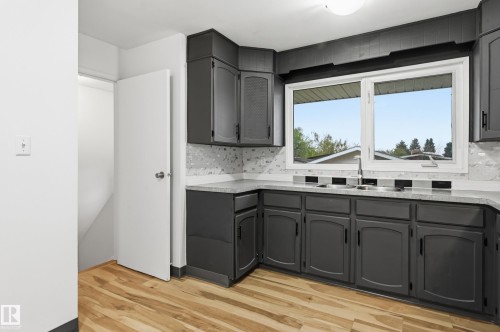 Kitchen featuring dark gray cabinetry, light wood-finish flooring, a double basin sink with a gooseneck faucet, light-toned countertops, and a patterned tile backsplash - 13903 87 Street, Edmonton, AB - Indoor Photo Showing Kitchen