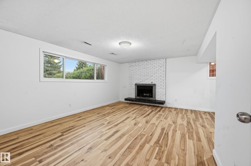 Spacious room featuring wood-finish flooring, a white brick fireplace with a dark hearth, and a large multi-pane window with exterior landscaping - 13903 87 Street, Edmonton, AB - Indoor Photo Showing Other Room With Fireplace