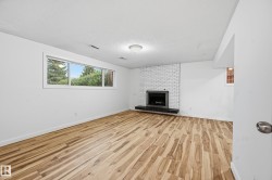 Spacious room featuring wood-finish flooring, a white brick fireplace with a dark hearth, and a large multi-pane window with exterior landscaping - 