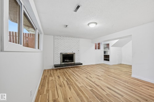 Spacious room featuring wood-finish flooring, a white brick fireplace with a dark hearth, built-in shelving, and white walls - 13903 87 Street, Edmonton, AB - Indoor With Fireplace