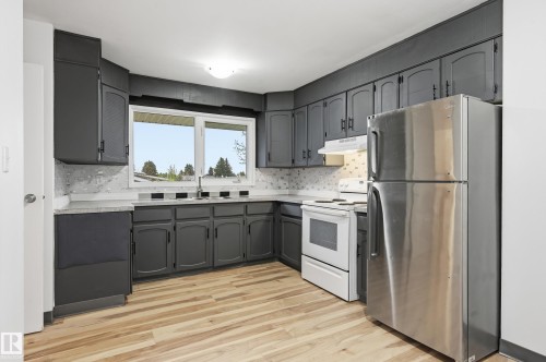 Kitchen with dark cabinetry, light-toned wood-finish flooring, a stainless steel refrigerator, white electric range, and a dual-basin sink under a large window - 13903 87 Street, Edmonton, AB - Indoor Photo Showing Kitchen With Stainless Steel Kitchen With Double Sink
