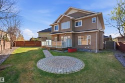 Expansive backyard featuring a circular paver patio, a raised wood-finish deck with glass panel railings, and a manicured lawn - 