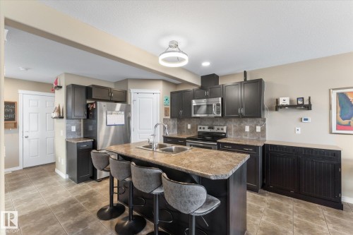 Kitchen featuring a central island with a double basin sink, dark wood cabinetry, stainless steel appliances, tile flooring, and a neutral backsplash - 21818 99A Avenue, Edmonton, AB - Indoor Photo Showing Kitchen With Double Sink