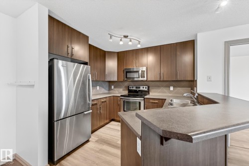Kitchen featuring wood-finish cabinetry, stainless steel appliances, a grey tile backsplash, and a breakfast bar with an undermount sink - 215 2208 44 Avenue, Edmonton, AB - Indoor Photo Showing Kitchen With Double Sink