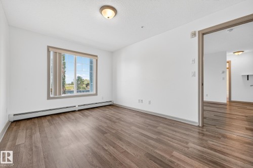 Spacious room featuring wood-finish flooring, a large window with blinds, and white baseboard heating - 215 2208 44 Avenue, Edmonton, AB - Indoor Photo Showing Other Room