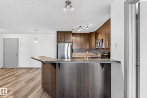 Open-concept kitchen featuring wood-finish flooring, dark wood cabinetry, stainless steel appliances, and a breakfast bar with a stone-finish countertop - 215 2208 44 Avenue, Edmonton, AB - Indoor Photo Showing Kitchen