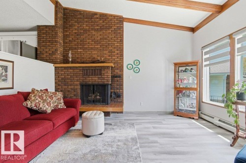 Spacious living area featuring a brick fireplace with a wood mantle, vaulted ceilings with exposed wood beams, large windows with horizontal blinds, and light wood-finish flooring - 8608 22 Ave, Edmonton, AB - Indoor Photo Showing Living Room With Fireplace