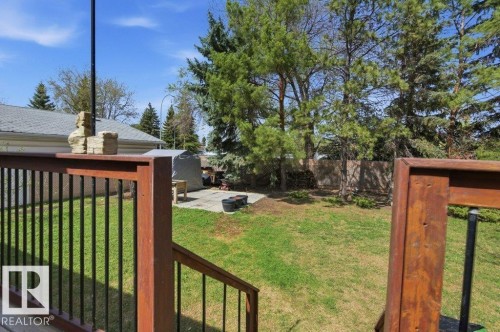 Spacious backyard featuring a wood deck with black metal balusters, concrete patio, mature evergreen trees, and a wooden perimeter fence - 8608 22 Ave, Edmonton, AB - Outdoor
