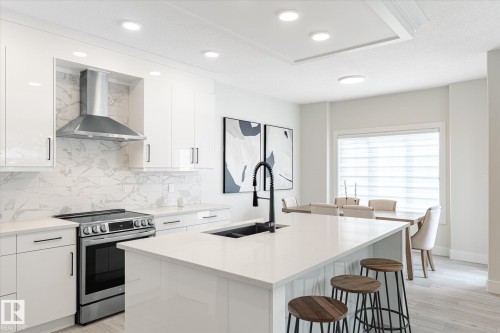 Modern kitchen featuring white cabinetry, a stainless steel range and hood vent, a large center island with a black faucet and undermount sink, and light wood-finish flooring - 1214 Eaton Lane, Edmonton, AB - Indoor Photo Showing Kitchen With Double Sink With Upgraded Kitchen
