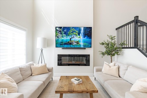 Bright living space featuring a wall-mounted television, an integrated linear electric fireplace, high ceilings, large windows, and a black metal spindle staircase - 1214 Eaton Lane, Edmonton, AB - Indoor Photo Showing Living Room With Fireplace