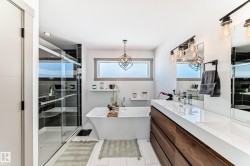 Modern bathroom featuring a freestanding soaking tub, a spacious glass-enclosed shower with dark tile surround, and a long wood-finish vanity with a white countertop - 