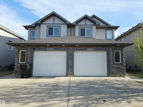 Contemporary duplex exterior featuring a stone and siding facade, two integrated single-car garages, an asphalt shingle roof, and multiple upper-level windows - 21321 94 Avenue, Edmonton, AB - Outdoor