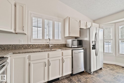 Kitchen featuring white cabinetry, speckled countertops, stainless steel appliances, and bay windows with plantation shutters - 18903 70 Avenue, Edmonton, AB - Indoor Photo Showing Kitchen
