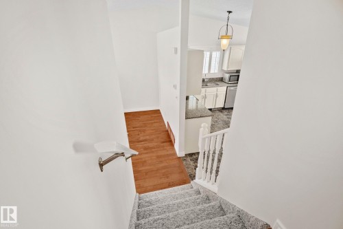 Carpeted staircase descending to a wood-finish flooring landing, with a kitchen featuring white cabinetry, light-toned countertops, and stainless steel appliances visible beyond a half-wall - 18903 70 Avenue, Edmonton, AB - Indoor Photo Showing Other Room