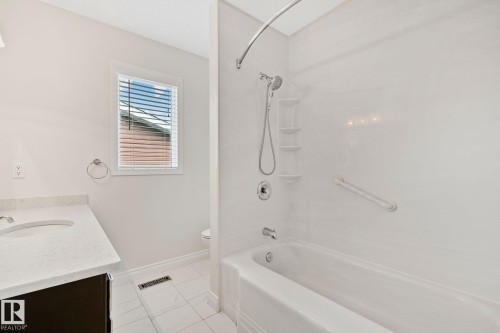 Bathroom featuring a white bathtub with a shower kit, white tile flooring, dark wood-finish vanity with a white countertop, and a white-framed window with blinds - 18903 70 Avenue, Edmonton, AB - Indoor Photo Showing Bathroom