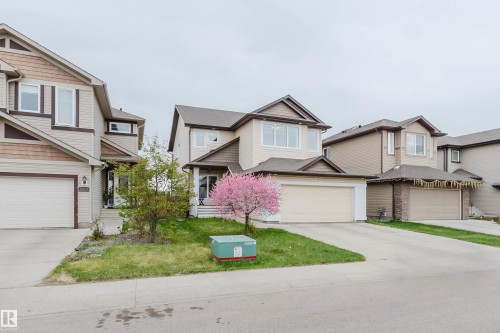 Two-story residence featuring light-toned siding with contrasting brown trim, an integrated garage, a paved driveway, and a front yard with a blooming tree - 17718 5A Avenue, Edmonton, AB - Outdoor With Facade