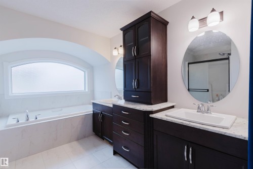 Bathroom featuring a built-in tub with marble-look surround, a large arched privacy window, dual vanity sinks with light countertops, dark wood-finish cabinetry, and circular mirrors with sconce lighting - 17718 5A Avenue, Edmonton, AB - Indoor Photo Showing Bathroom