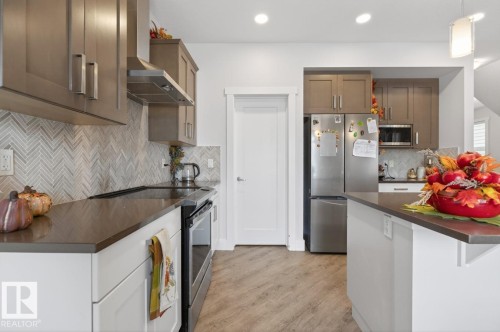 Kitchen featuring neutral shaker-style cabinetry, a herringbone tile backsplash, stainless steel appliances, dark countertops, and wood-finish flooring - 7942 Erasmus Crescent, Edmonton, AB - Indoor Photo Showing Kitchen With Upgraded Kitchen