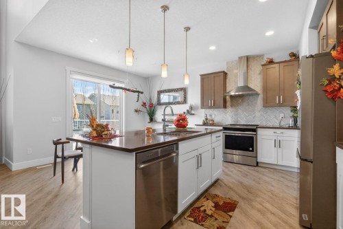 Kitchen featuring a central island with integrated dishwasher, stainless steel appliances, two-tone cabinetry, herringbone tile backsplash, and wood-finish flooring - 7942 Erasmus Crescent, Edmonton, AB - Indoor Photo Showing Kitchen With Upgraded Kitchen