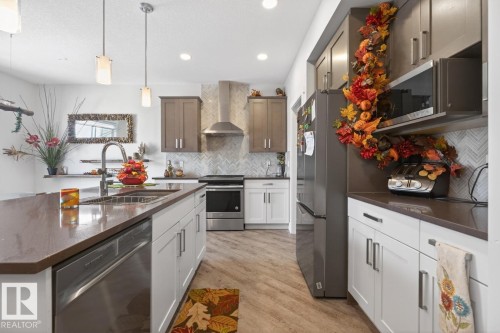 Modern kitchen featuring an expansive center island with an integrated stainless steel sink and dishwasher, pendant lighting, shaker-style cabinetry, and wood-finish flooring - 7942 Erasmus Crescent, Edmonton, AB - Indoor Photo Showing Kitchen With Double Sink With Upgraded Kitchen