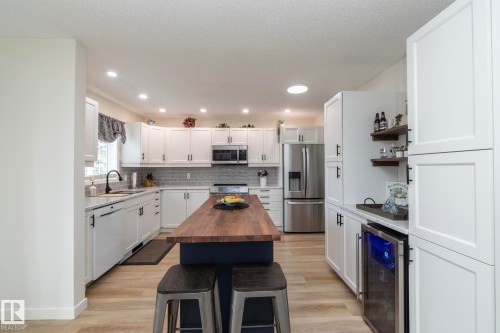 Modern kitchen featuring a central island with wood-finish countertop, stainless steel appliances, white cabinetry, light wood-finish flooring, and a beverage refrigerator - 25 1904 Mill Woods Road Road, Edmonton, AB - Indoor Photo Showing Kitchen With Upgraded Kitchen