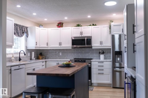 Modern kitchen featuring a central island with a wood-finish countertop, white cabinetry with black hardware, stainless steel appliances, a subway tile backsplash, and recessed lighting - 25 1904 Mill Woods Road Road, Edmonton, AB - Indoor Photo Showing Kitchen With Upgraded Kitchen