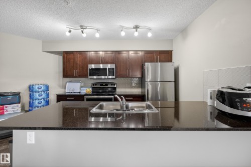 Kitchen featuring dark wood-finish cabinetry, stainless steel appliances, a dark countertop, and track lighting fixtures - 314 3211 James Mowatt Trail Sw, Edmonton, AB - Indoor Photo Showing Kitchen With Stainless Steel Kitchen With Double Sink