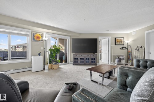 Carpeted living area featuring a large window, sliding glass door, white baseboards, and neutral wall tones - 314 3211 James Mowatt Trail Sw, Edmonton, AB - Indoor Photo Showing Living Room