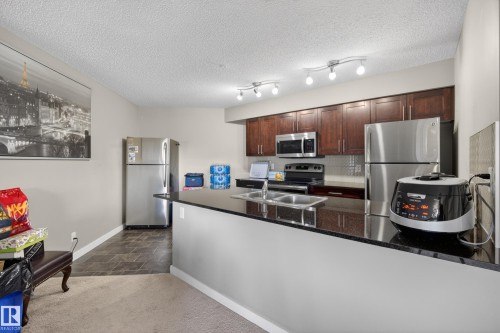 Kitchen featuring dark wood-finish cabinetry, black countertops, and stainless steel appliances - 314 3211 James Mowatt Trail Sw, Edmonton, AB - Indoor Photo Showing Kitchen With Stainless Steel Kitchen With Double Sink