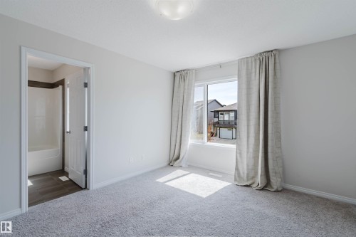Carpeted room featuring a large window with drapes, a flush mount ceiling light, and an open doorway revealing a bathroom with a white tub and tiled walls - 2082 Price Landing, Edmonton, AB - Indoor