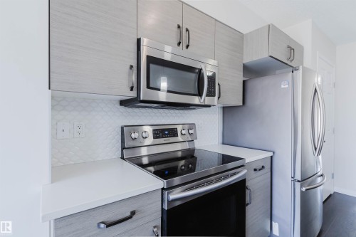 Modern kitchen featuring light gray wood-finish cabinetry, stainless steel appliances, white countertop surfaces, and a white scallop-patterned tile backsplash - 2082 Price Landing, Edmonton, AB - Indoor Photo Showing Kitchen With Stainless Steel Kitchen